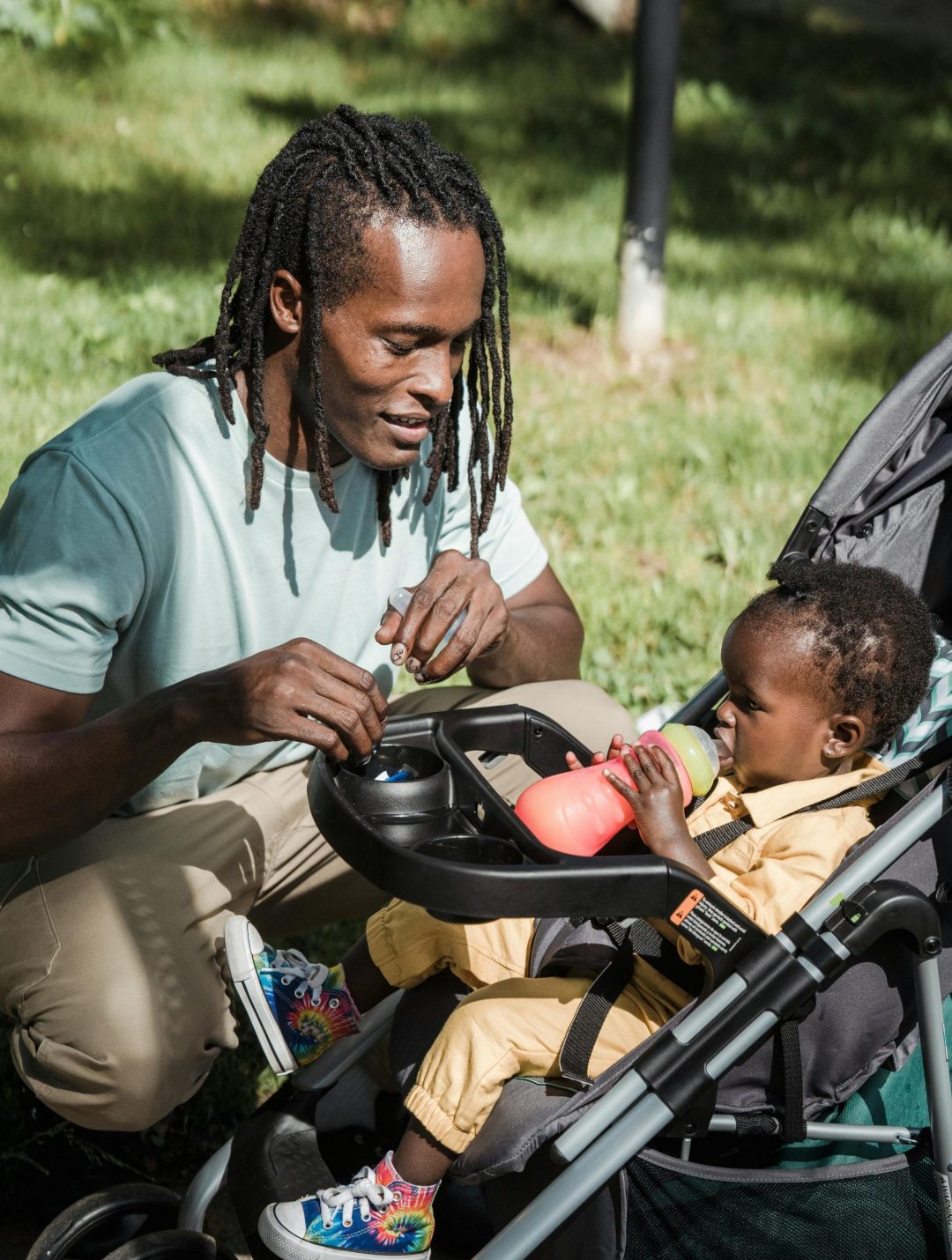 A father lovingly feeds his toddler in a stroller during a sunny day outdoors, highlighting family bonding.
